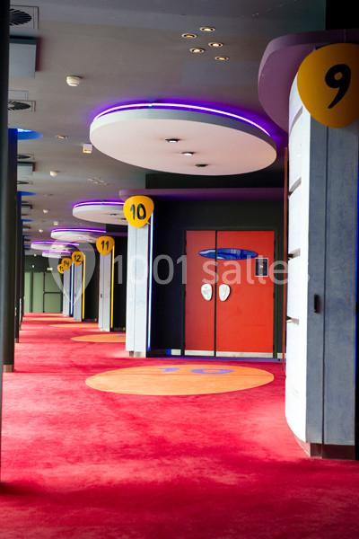 Location salle Ivry-sur-Seine (Val-de-Marne) - Pathé Quai D'ivry #6 Couloir d'un cinéma avec moquette rouge, portes numérotées et éclairage violet au plafond.