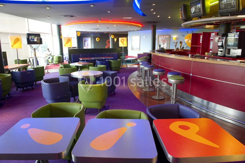 Location salle Ivry-sur-Seine (Val-de-Marne) - Pathé Quai D'ivry #4 Salle colorée avec tables décorées, fauteuils, bar rouge et écrans suspendus dans un espace moderne et lumineux.