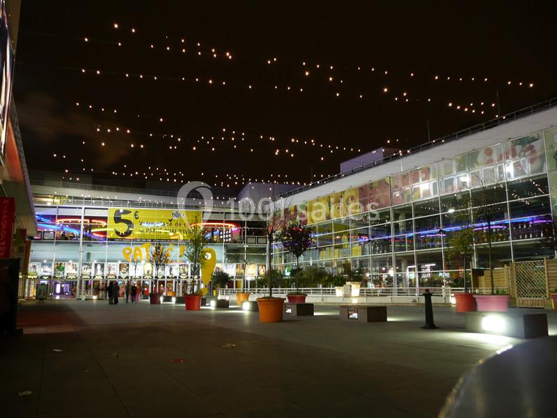 Location salle Ivry-sur-Seine (Val-de-Marne) - Pathé Quai D'ivry #2 Cour extérieure illuminée la nuit, entourée de bâtiments vitrés avec des affiches colorées et des guirlandes lumineuses.