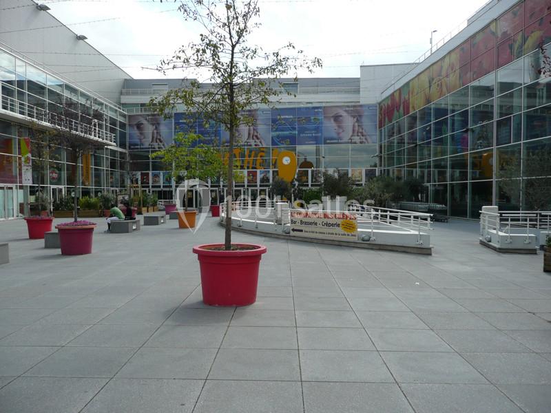 Location salle Ivry-sur-Seine (Val-de-Marne) - Pathé Quai D'ivry #7 Cour extérieure d'un centre commercial avec des pots de fleurs colorés, des arbres et une façade vitrée en arrière-plan.