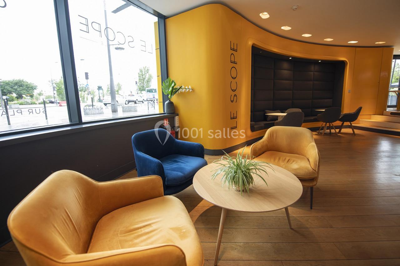 Espace d'attente moderne avec fauteuils colorés, table basse et murs jaunes, éclairé par de grandes fenêtres.