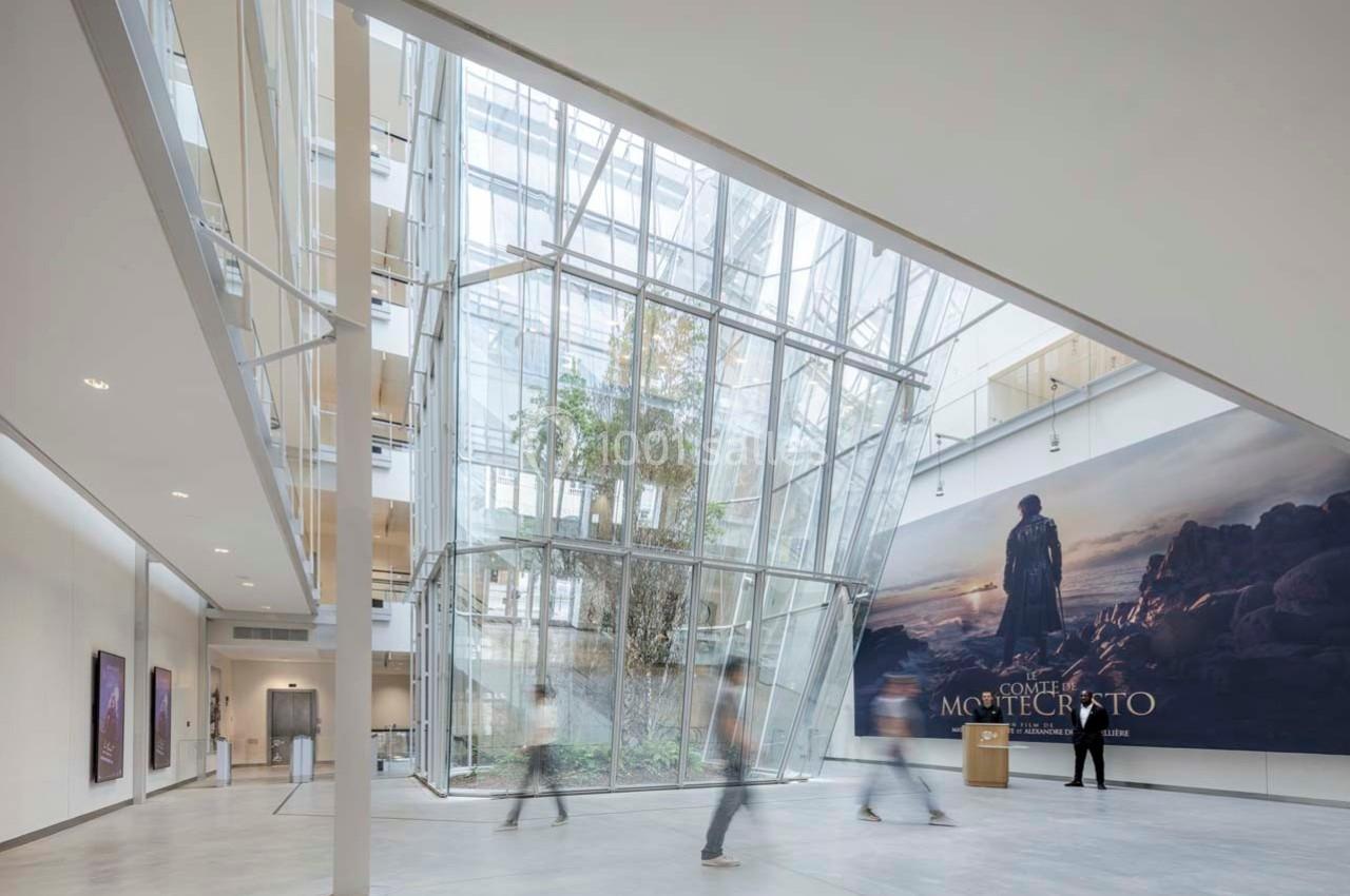 Location salle Paris 9 (Paris) - Pathé Palace  #3 Hall lumineux avec verrière inclinée, plantes au centre et affiche murale représentant un paysage rocheux.