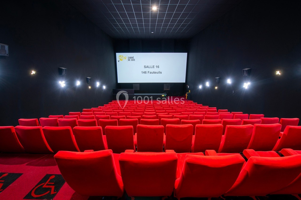 Salle de cinéma avec des rangées de fauteuils rouges vides, écran blanc et éclairage tamisé.