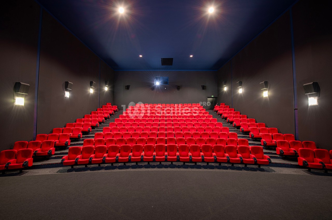 Salle de cinéma vide avec des rangées de fauteuils rouges, écran éteint et éclairage tamisé.