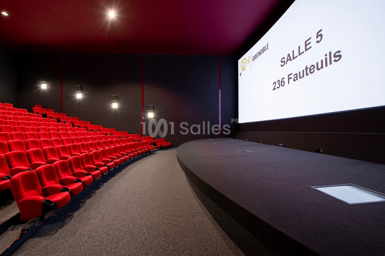 Salle de cinéma vide avec écran blanc affichant ’Salle 5, 236 fauteuils’ et rangées de sièges rouges.