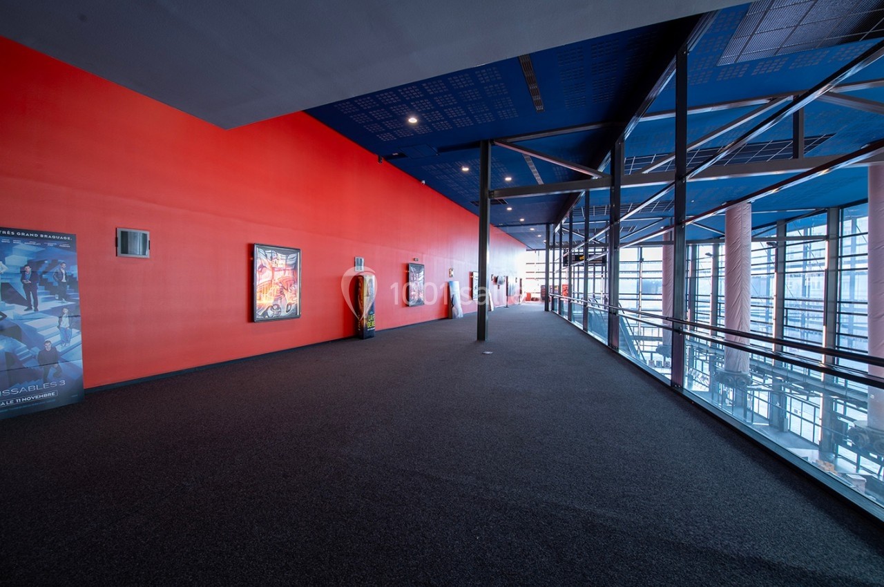 Couloir spacieux avec mur rouge, affiches encadrées, sol en moquette sombre et grandes baies vitrées à droite.