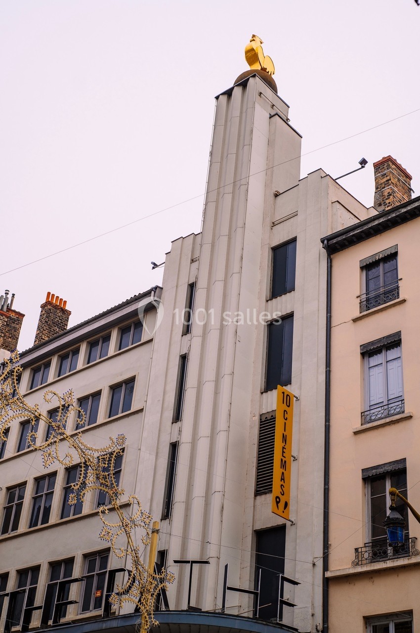 Façade d'un cinéma art déco avec une sculpture de coq doré au sommet, entourée de bâtiments urbains.