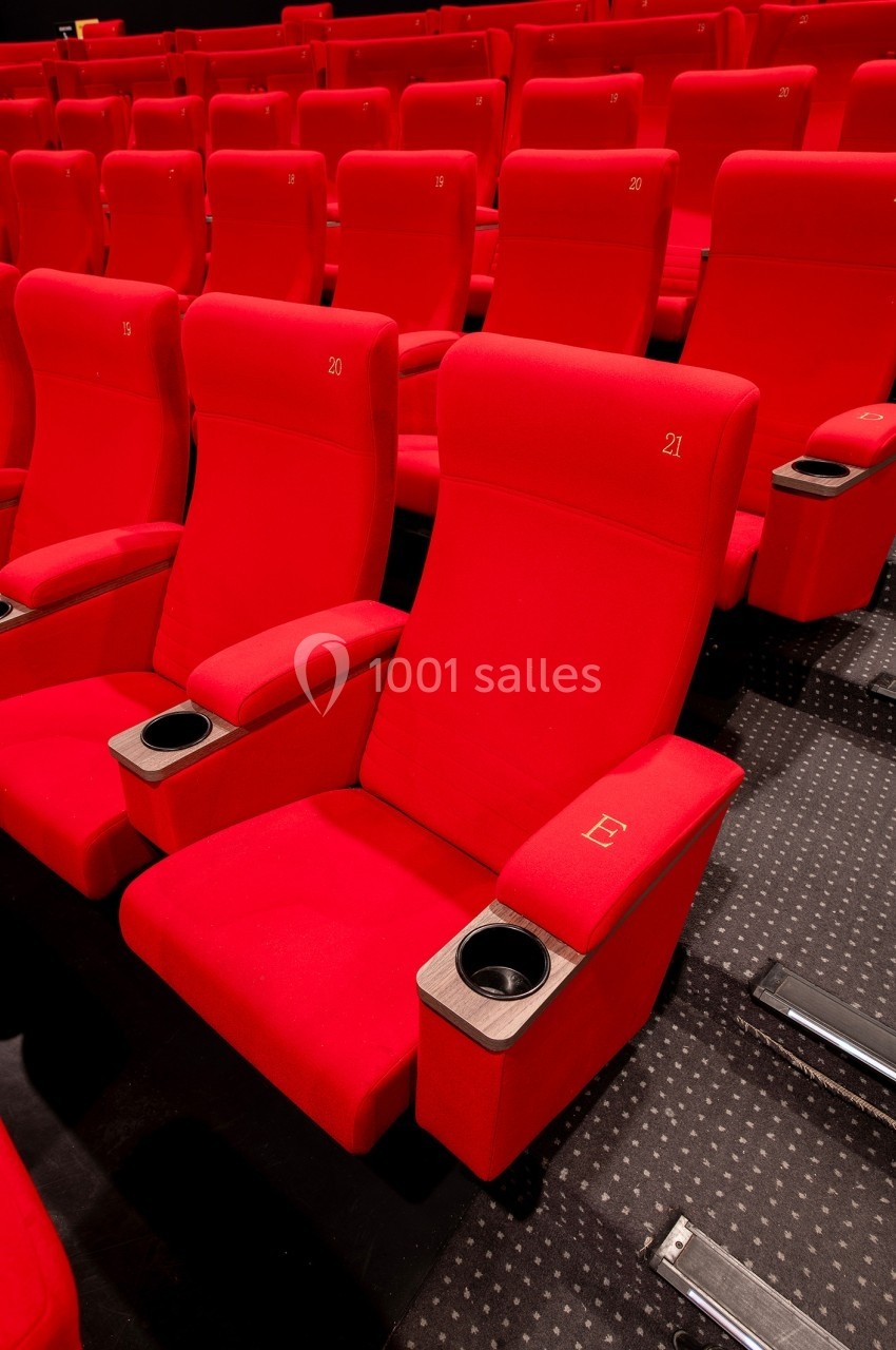 Rangée de fauteuils de cinéma rouges avec accoudoirs et porte-gobelets, sur un sol en moquette sombre.