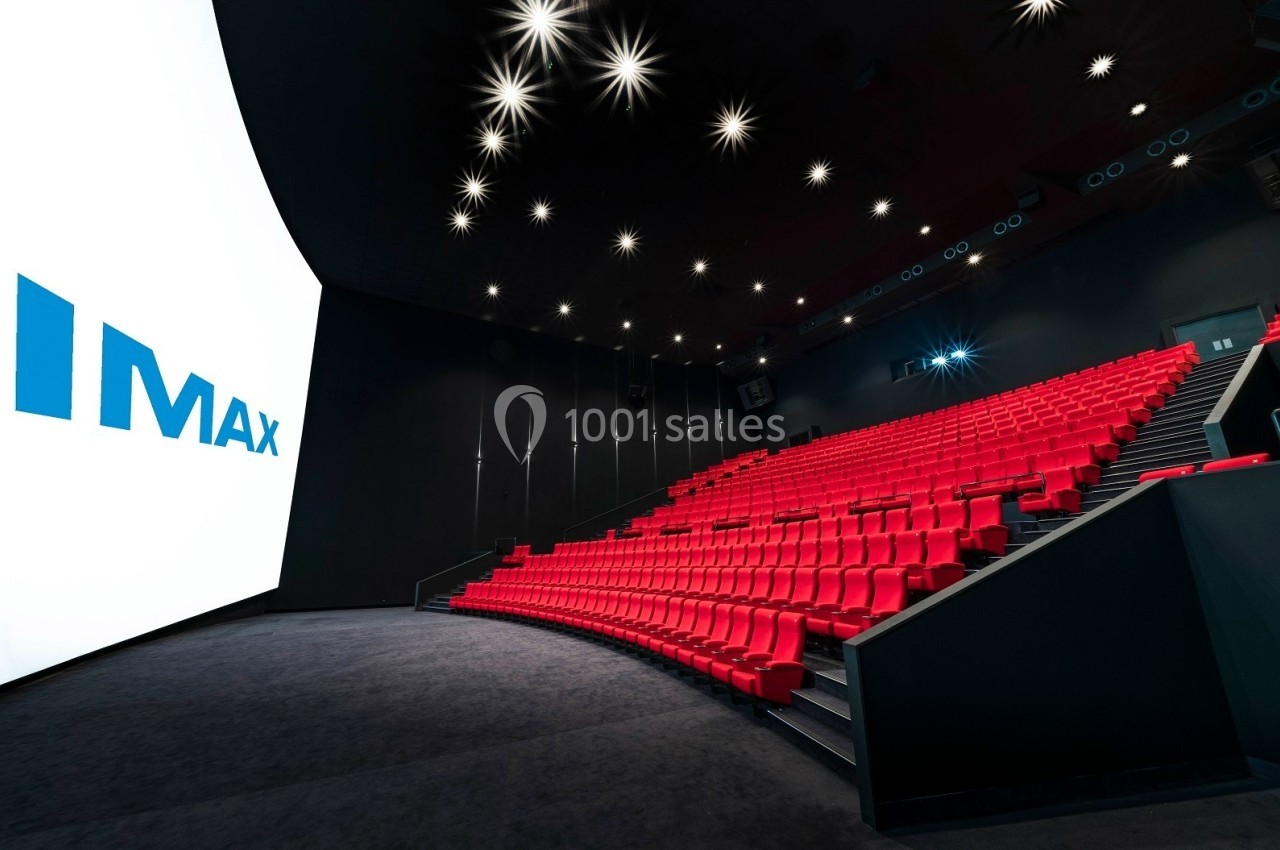 Salle de cinéma IMAX avec des rangées de sièges rouges, un grand écran blanc et un éclairage au plafond.