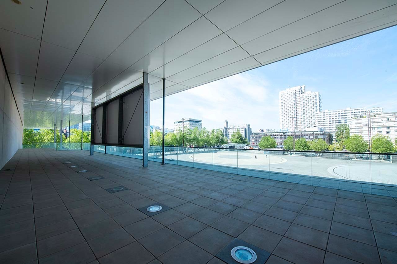 Vue d'une terrasse couverte avec sol carrelé, balustrade en verre et panorama urbain sous un ciel dégagé.