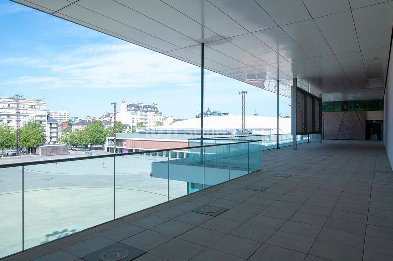 Vue depuis une terrasse couverte avec garde-corps en verre, donnant sur une place urbaine et des bâtiments modernes.