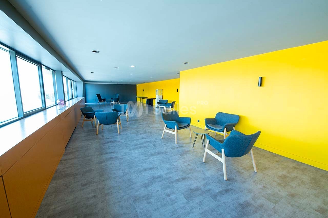 Salle lumineuse avec murs jaunes, chaises bleues, tables basses en verre et grandes fenêtres laissant entrer la lumière…
