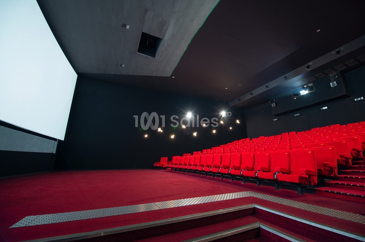 Salle de cinéma vide avec des rangées de fauteuils rouges, un écran blanc et un éclairage tamisé.