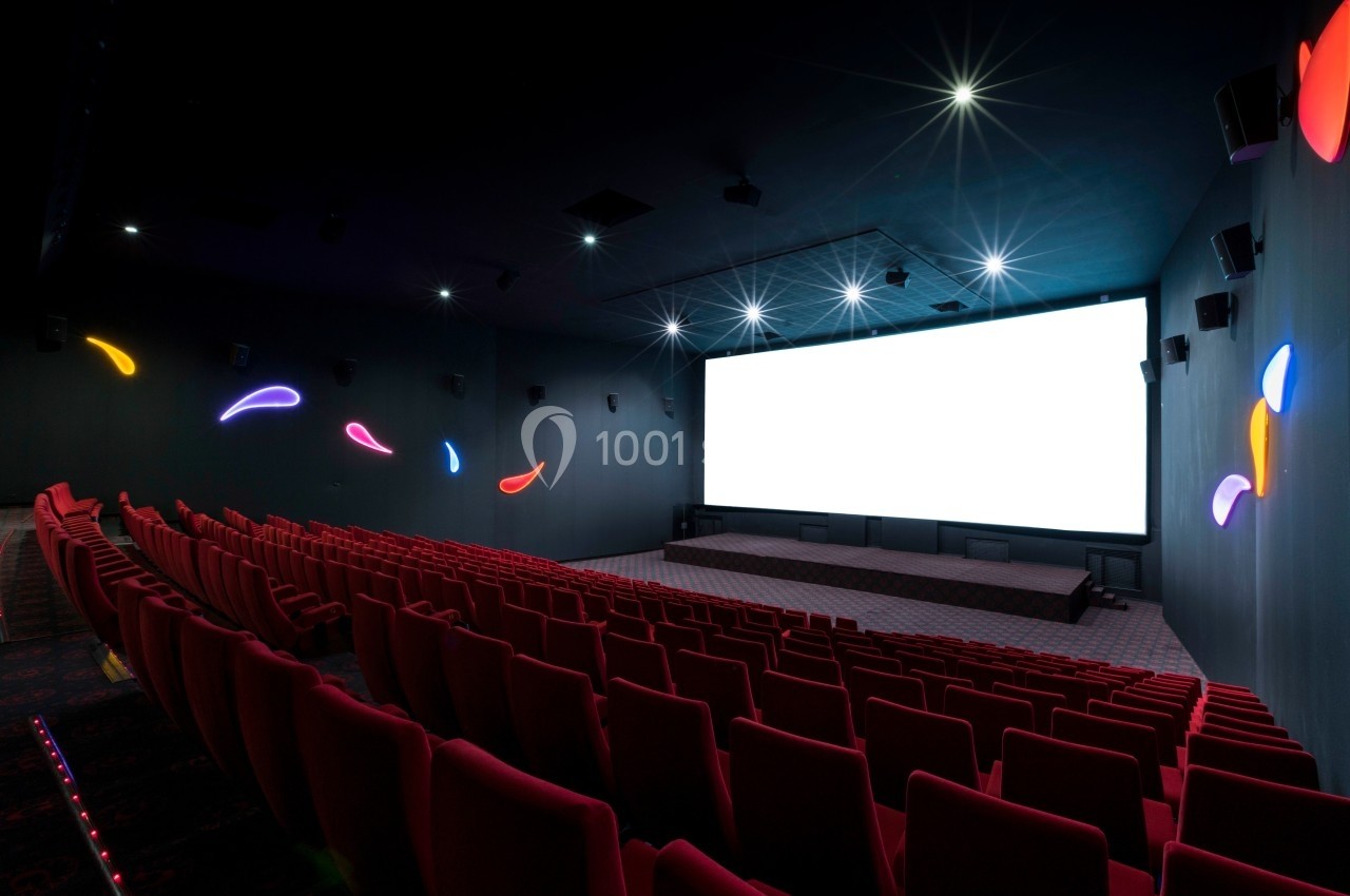 Salle de cinéma vide avec des rangées de fauteuils rouges, un grand écran blanc et des lumières décoratives colorées.