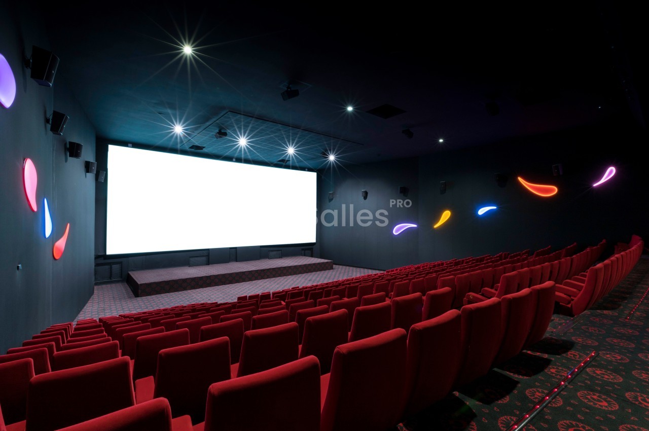 Salle de cinéma vide avec des rangées de sièges rouges, un grand écran blanc et des lumières décoratives colorées.