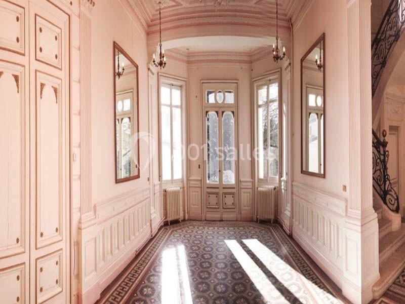 Salle lumineuse avec murs roses, grandes fenêtres, moulures décoratives, sol en carreaux et escalier en fer forgé visible.