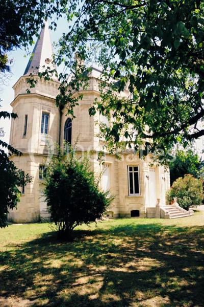 Façade d'un petit château en pierre entouré d'arbres et d'un jardin ombragé par une journée ensoleillée.