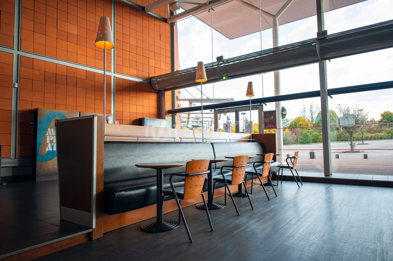 Intérieur d'un café moderne avec des tables, des chaises en bois et de grandes baies vitrées donnant sur l'extérieur.