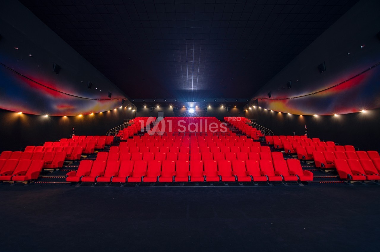 Salle de cinéma vide avec des rangées de sièges rouges et un écran illuminé au fond.