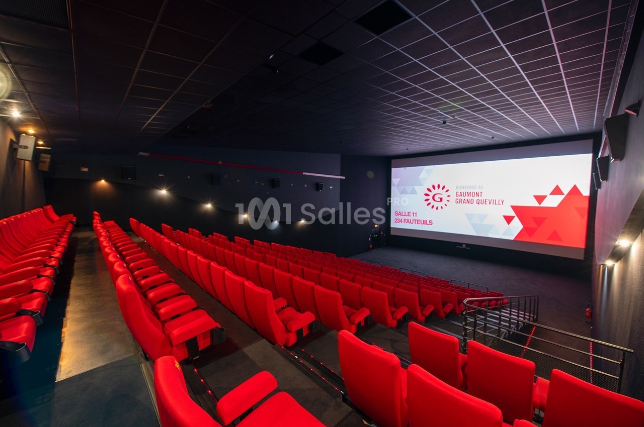 Salle de cinéma vide avec rangées de sièges rouges et grand écran affichant un visuel promotionnel.