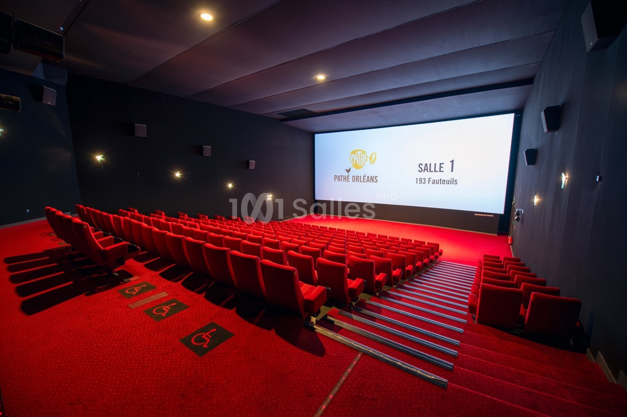 Salle de cinéma vide avec rangées de sièges rouges, écran blanc affichant ’Salle 1’ et signalétique d'accessibilité au sol.