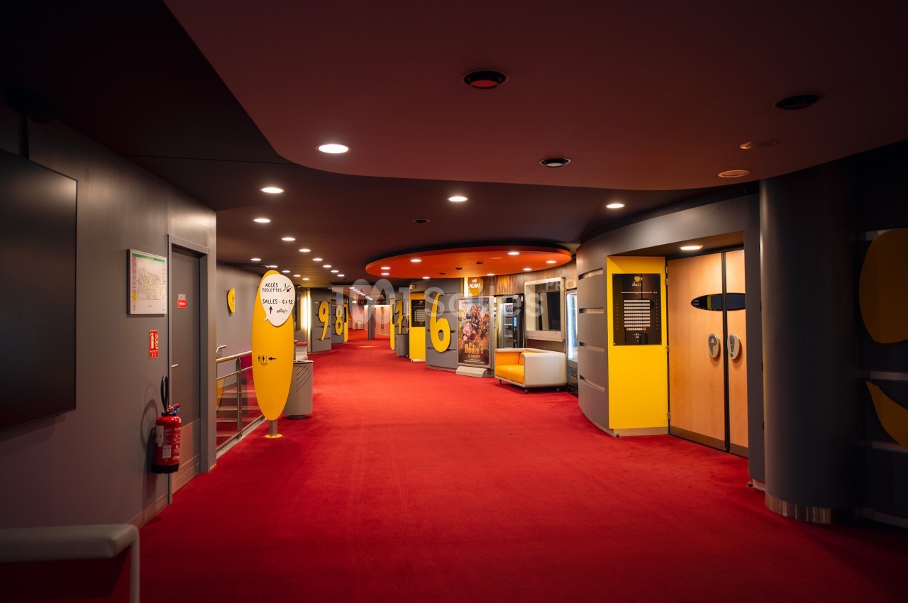 Couloir d'un cinéma avec moquette rouge, numéros de salles jaunes et affiches de films sur les murs.