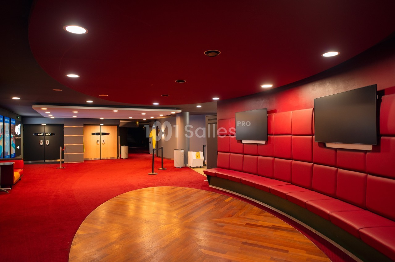 Hall d'entrée moderne avec sièges rouges, écrans muraux et sol en bois, éclairé par des spots au plafond.