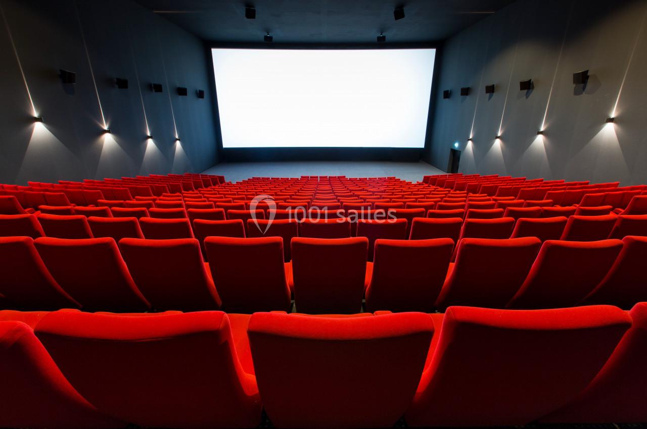 Salle de cinéma vide avec des rangées de fauteuils rouges et un grand écran blanc allumé à l'arrière-plan.