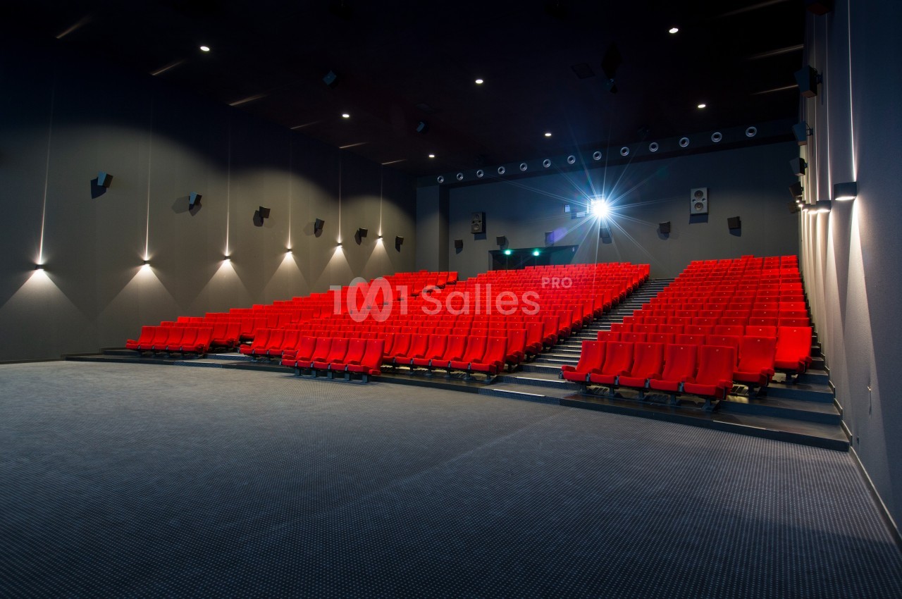 Salle de cinéma vide avec sièges rouges alignés et écran éteint, éclairée par des lumières murales et un projecteur.