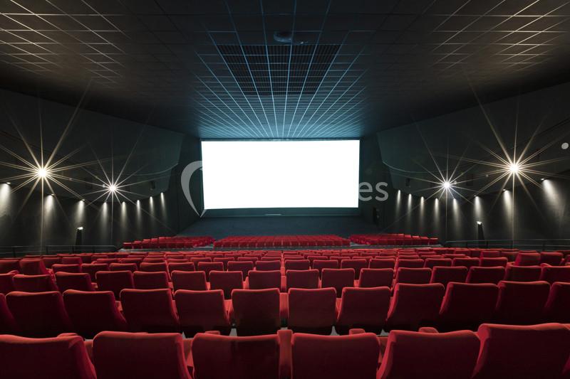 Salle de cinéma vide avec des rangées de sièges rouges et un grand écran blanc allumé au fond.