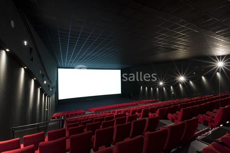 Salle de cinéma vide avec des rangées de sièges rouges et un grand écran blanc allumé.