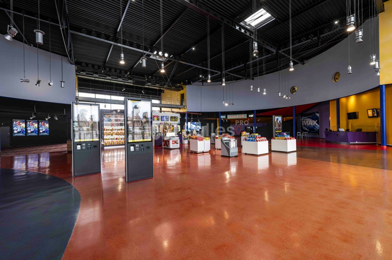 Hall spacieux d'un cinéma avec sol rouge, vitrines d'exposition, comptoirs de confiseries et affiches de films.
