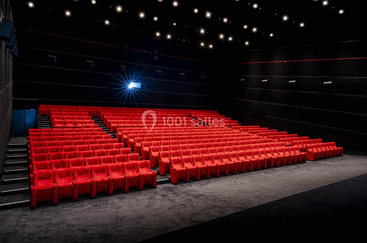 Salle de cinéma vide avec des rangées de fauteuils rouges et un projecteur allumé.