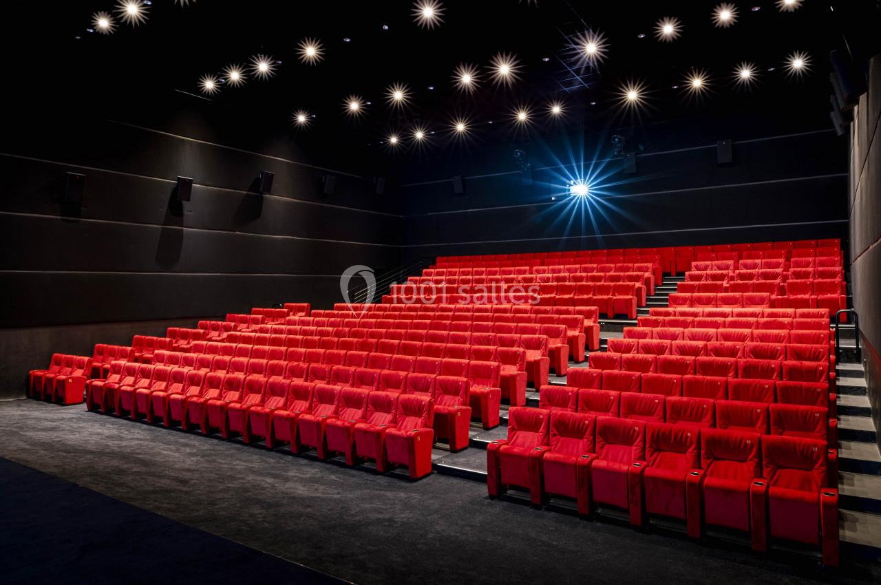 Salle de cinéma vide avec des rangées de fauteuils rouges et un projecteur allumé.