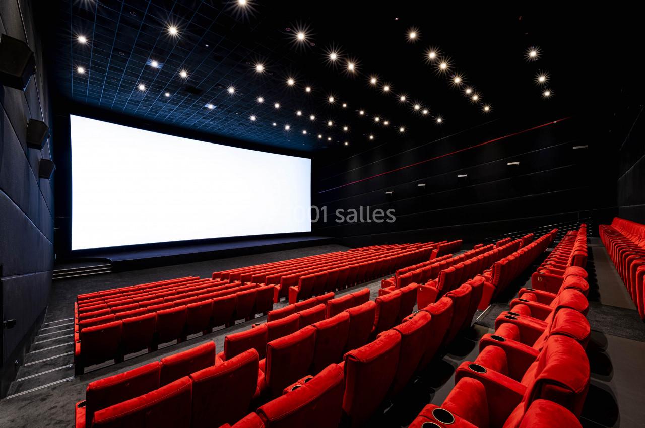 Salle de cinéma vide avec des rangées de fauteuils rouges et un grand écran blanc allumé.