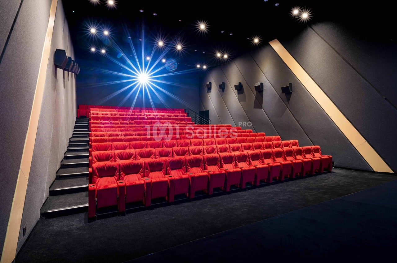 Salle de cinéma vide avec des rangées de sièges rouges, éclairée par un projecteur lumineux au plafond.