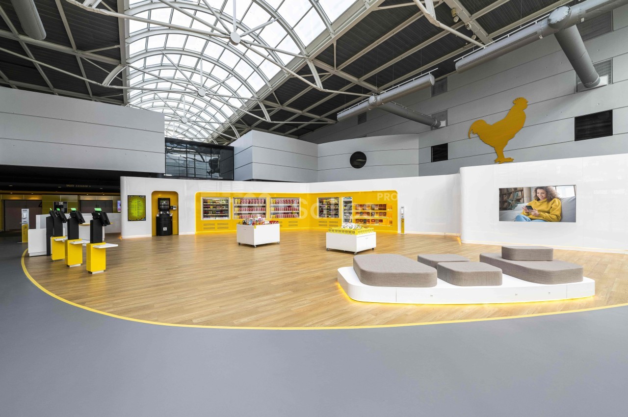 Vue d'un espace intérieur moderne avec bornes interactives, étagères jaunes et une grande sculpture de poule.