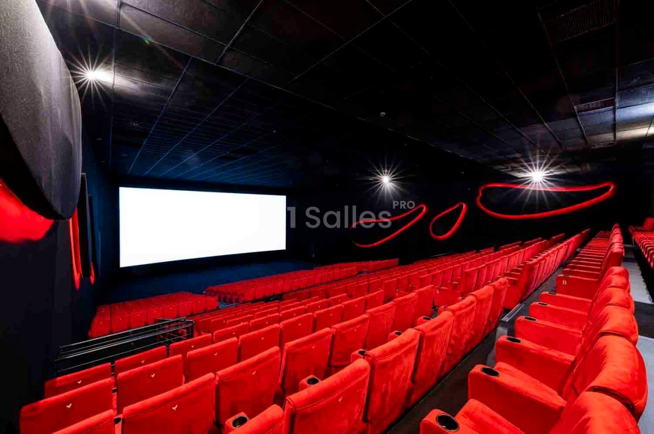 Salle de cinéma vide avec sièges rouges, écran blanc allumé et éclairage tamisé.