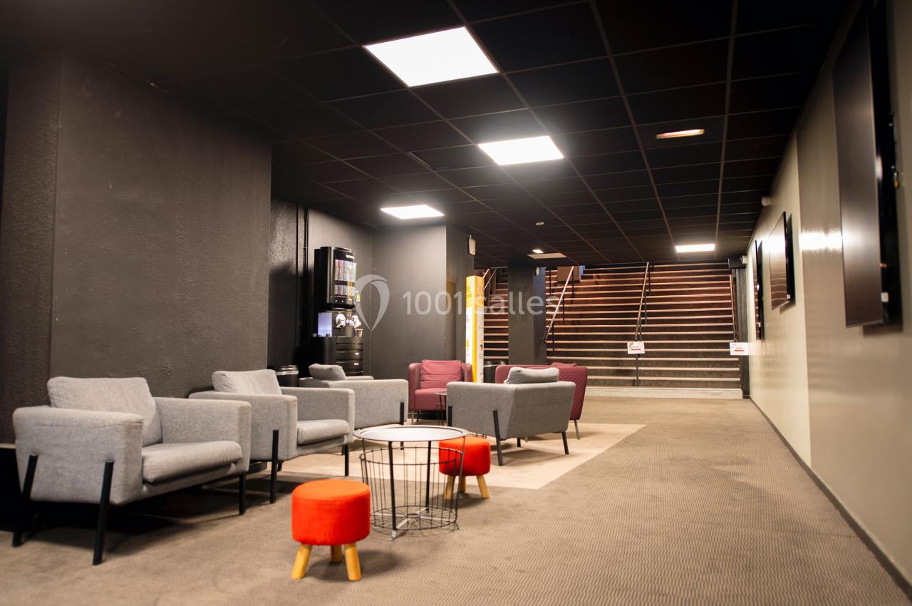 Espace d'attente avec fauteuils gris, tabourets colorés et escalier en arrière-plan, éclairé par des plafonniers.
