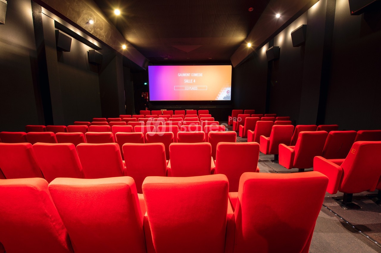 Salle de cinéma vide avec des sièges rouges alignés face à un écran de projection allumé.