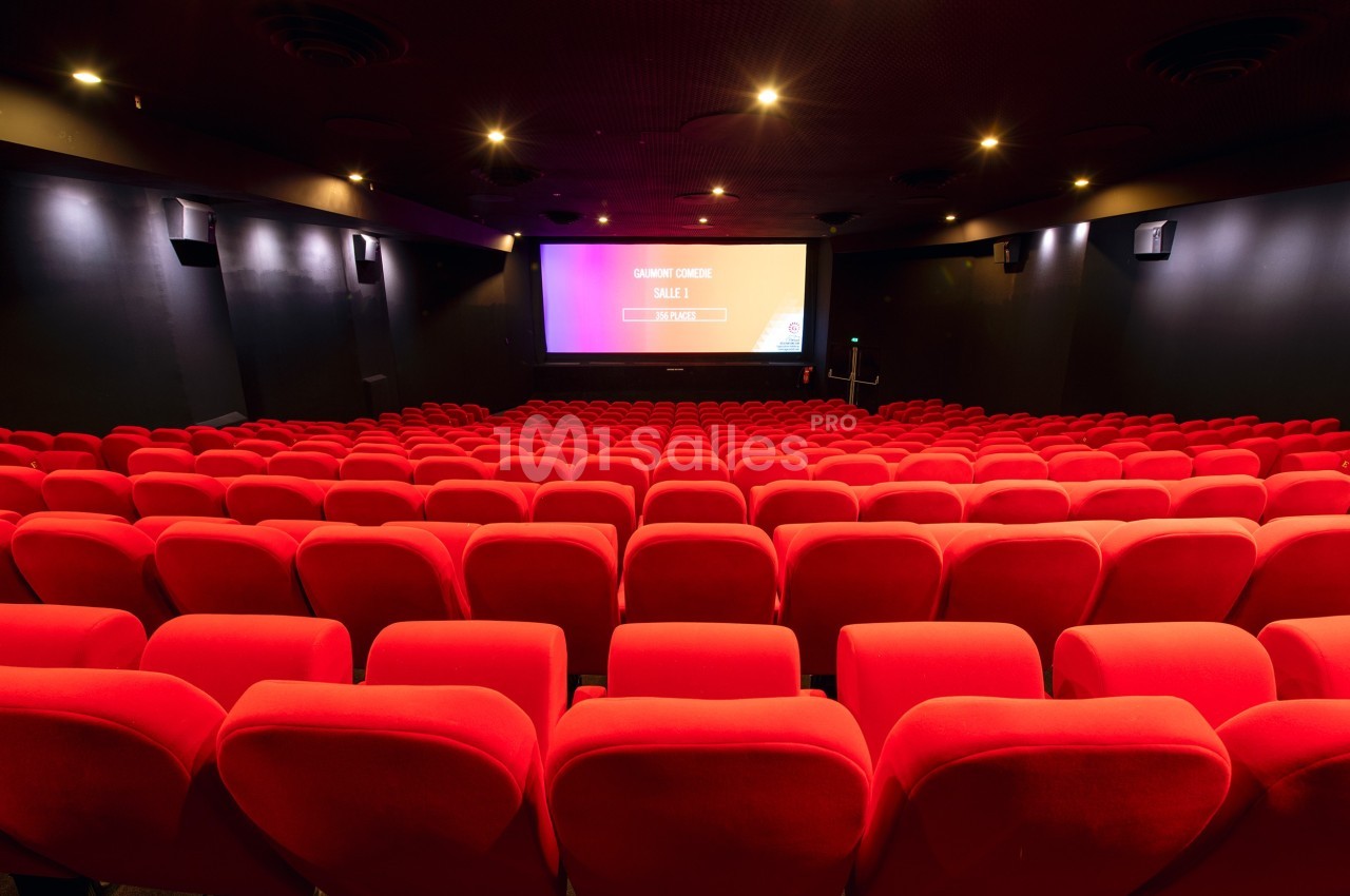 Salle de cinéma vide avec des rangées de fauteuils rouges et un écran allumé au fond.