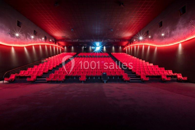 Salle de cinéma vide avec rangées de sièges rouges, éclairage tamisé et écran allumé au fond.