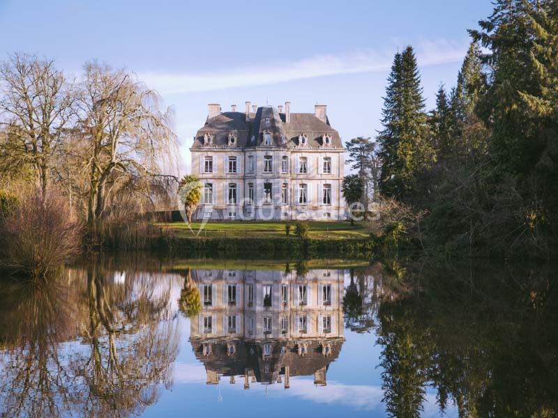 Façade d'un château entouré d'arbres, reflété dans un étang calme sous un ciel dégagé.