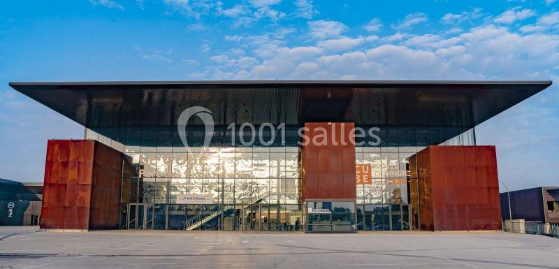 Façade moderne d'un bâtiment avec des murs en verre et acier rouillé, sous un ciel partiellement nuageux.