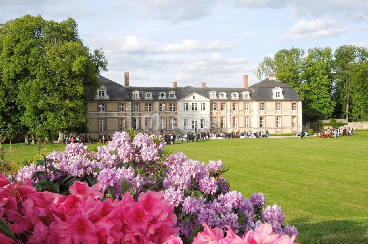 Façade d'un château entouré de pelouses et de fleurs roses au premier plan, sous un ciel partiellement nuageux.