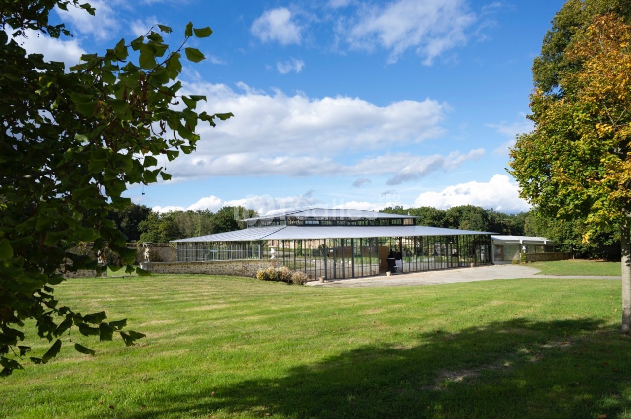 Bâtiment moderne en verre et métal entouré de pelouses et d'arbres sous un ciel partiellement nuageux.