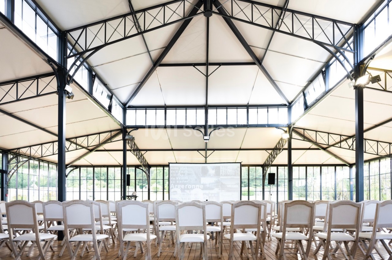 Salle lumineuse avec charpente métallique, rangées de chaises blanches alignées face à un écran de projection.