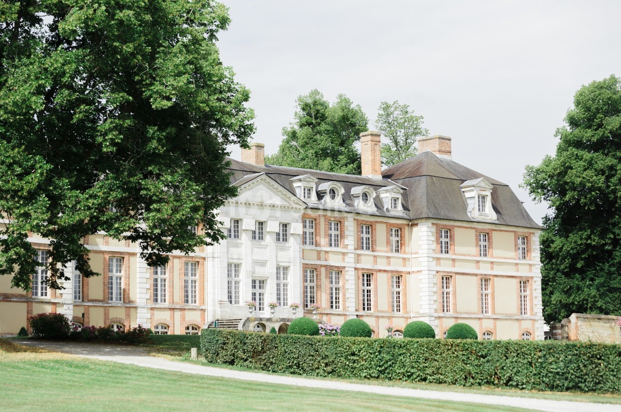 Façade d'un château en pierre et briques entouré de pelouses et d'arbres sous un ciel clair.