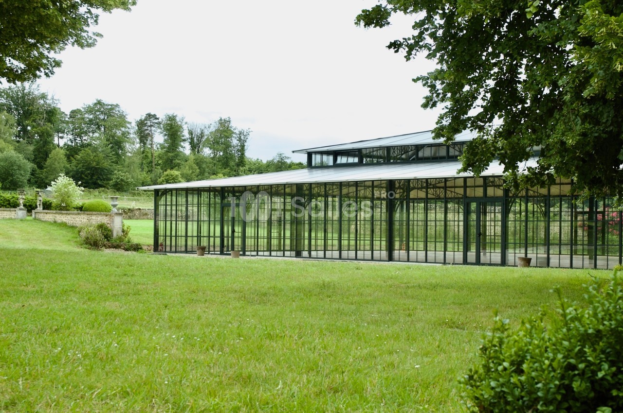 Serre en verre entourée de pelouses et d'arbres dans un cadre naturel verdoyant.