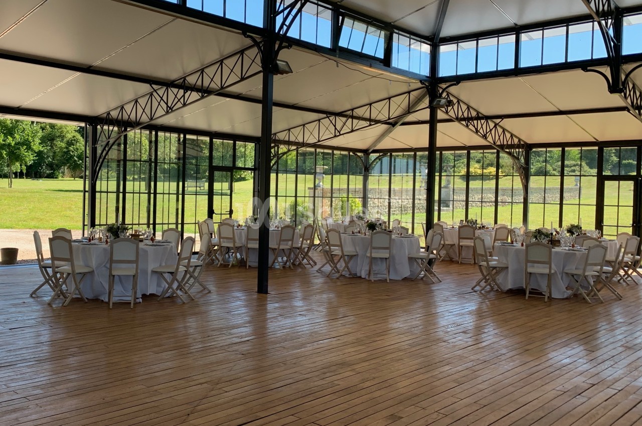 Salle lumineuse avec parquet en bois, tables rondes dressées et grandes baies vitrées donnant sur un espace vert.